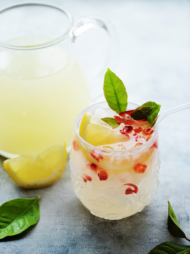 Lemonade in glass garnished with lemon & strawberries. Jar of lemonade in background