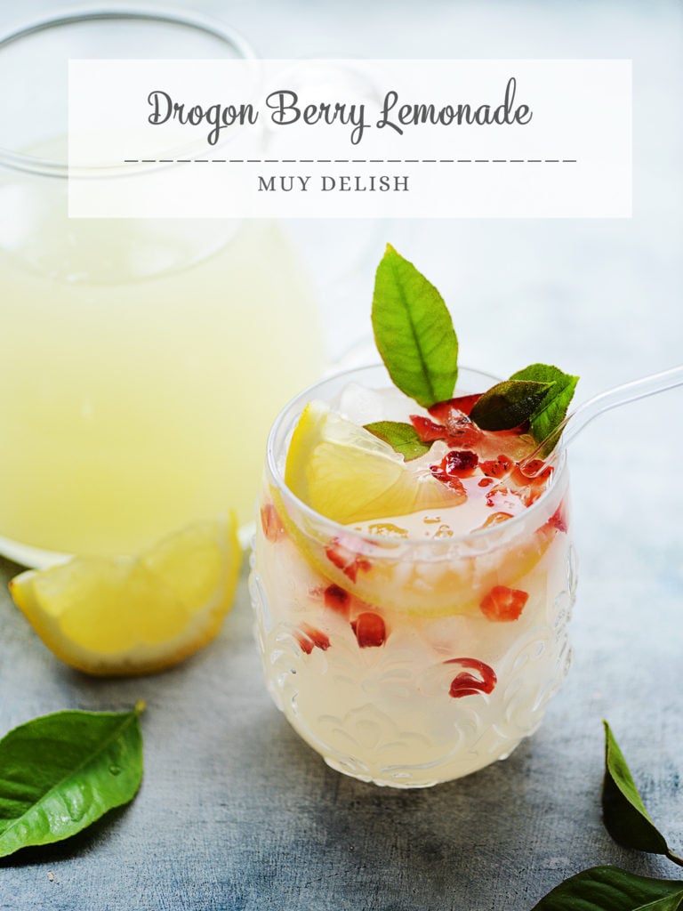 Lemonade in glass garnished with lemon & strawberries. Jar of lemonade in background