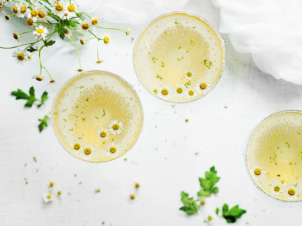 An overhead shot of three glasses with cocktail mix. A bouquet of chamomile flowers on the top left corner. 