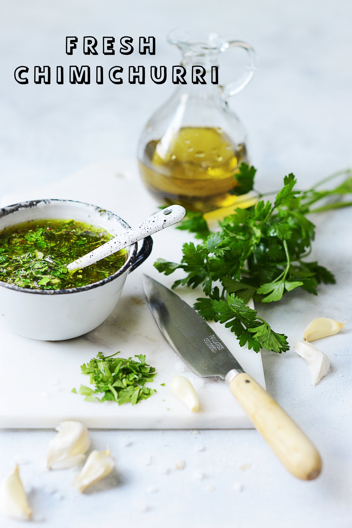 A white cutting board with a white cup filled with chimichurri. A bunch of herbs on the side
