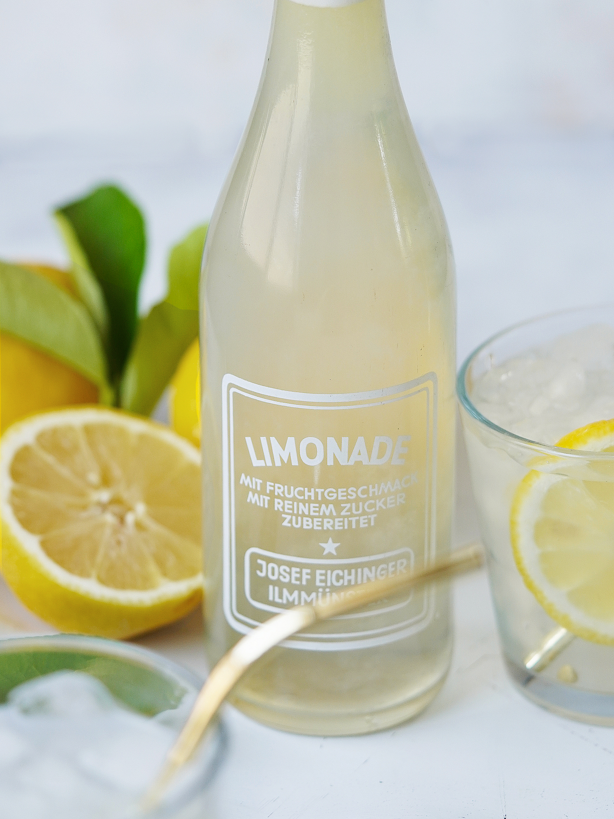 A bottle of limonada with lemons on the side and a glass with ice.