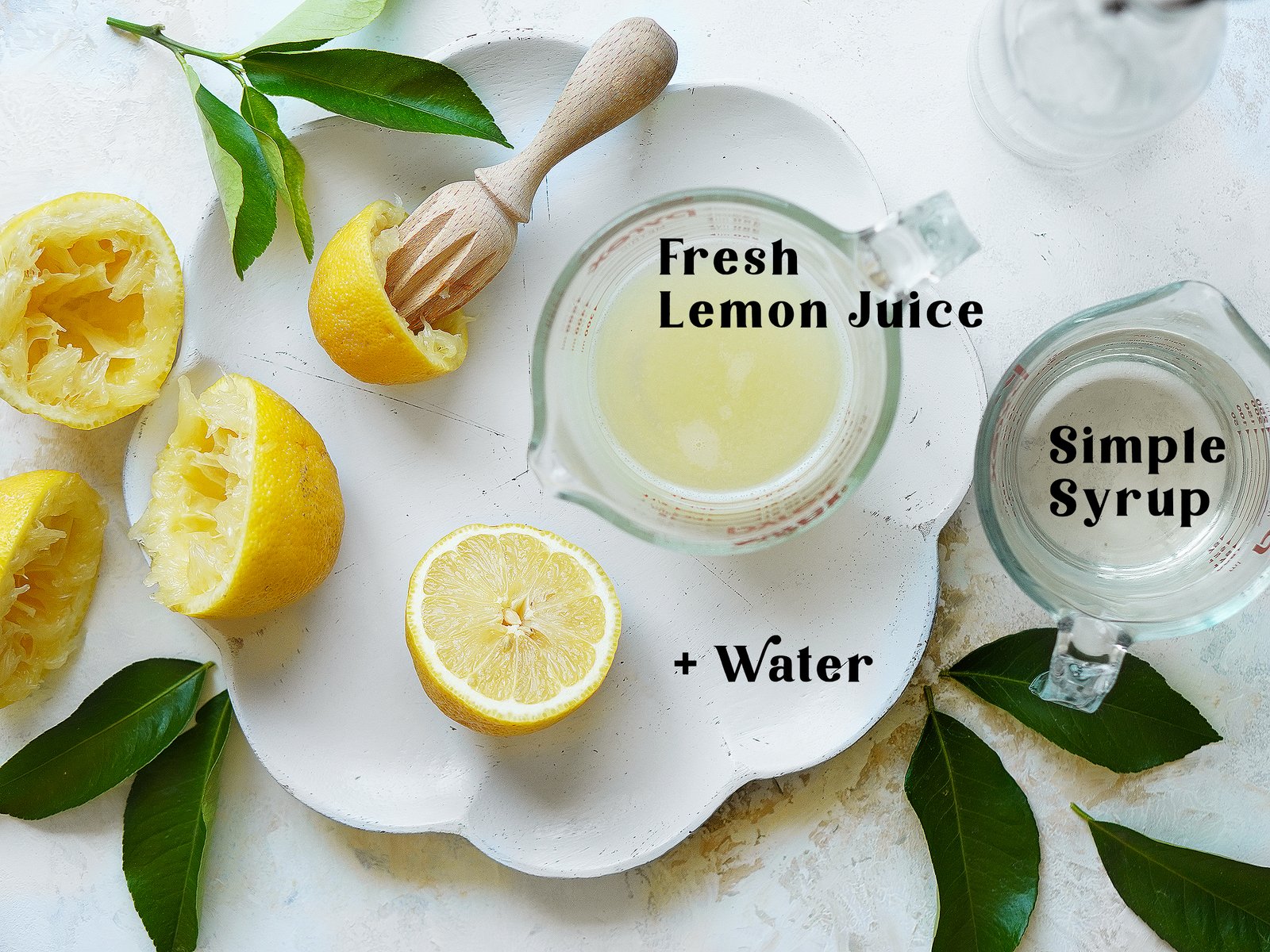 A white serving plate with fresh squeezed lemons, a cup with lemon juice and a cup with simple syrup