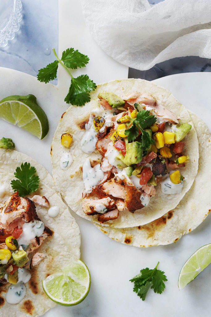 Two Salmon Tacos on a small white board with limes on the side