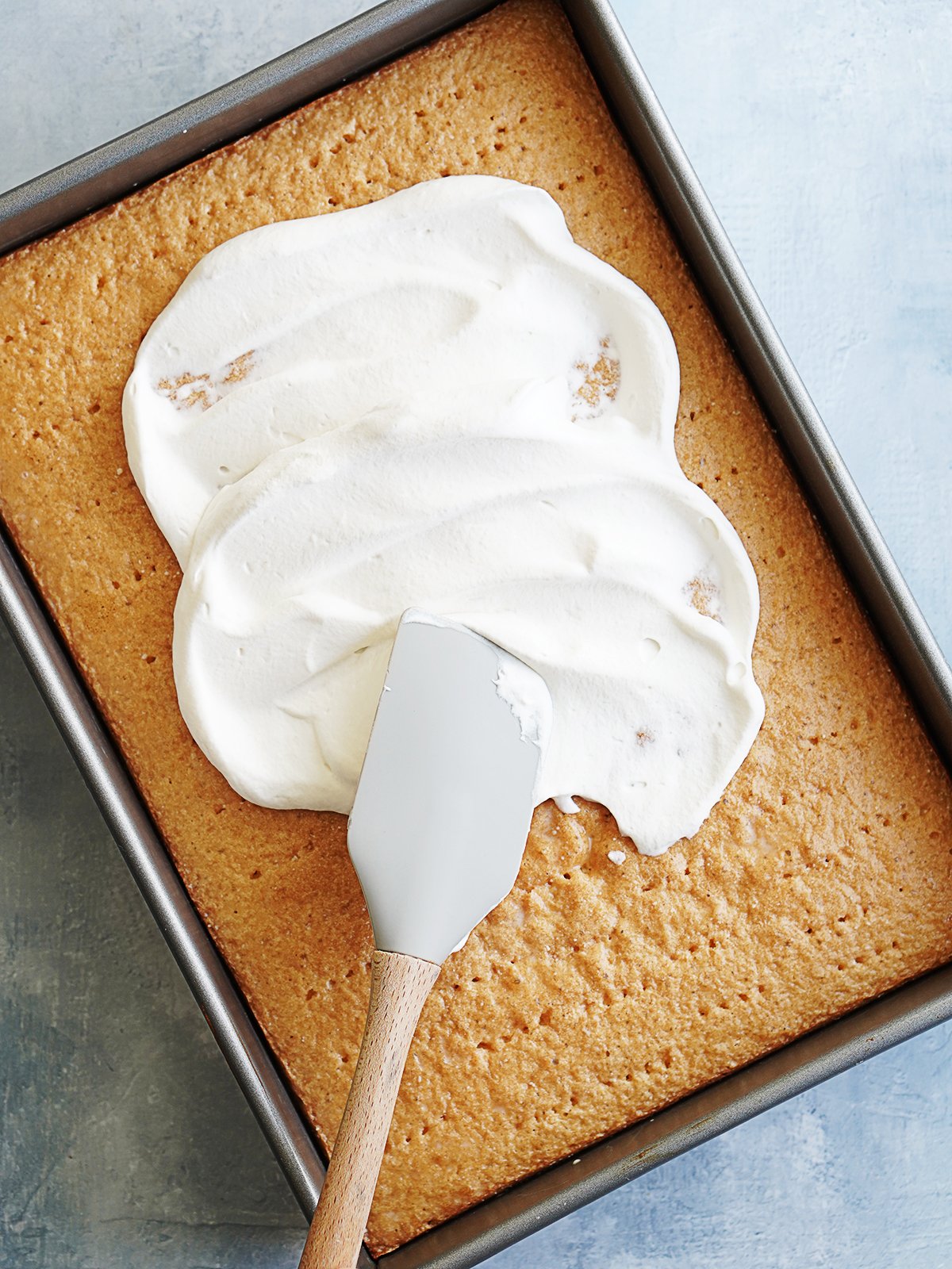 Cake in a baking pan being frosted with whipped cream