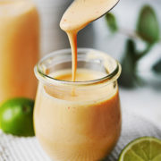 A spoon drizzling creamy, light orange Chipotle Crema into a small glass jar, with a lime and lime wedge nearby on a white textured plate. A bottle of the same dressing is in the background.