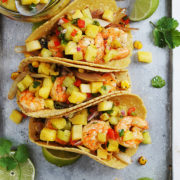 Three Tacos De Camaron folded on a metal tray with pineapple in a bowl on the side.