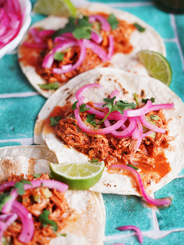 3 tacos of cochinita pibil topped with red pickled onions and limes on the side