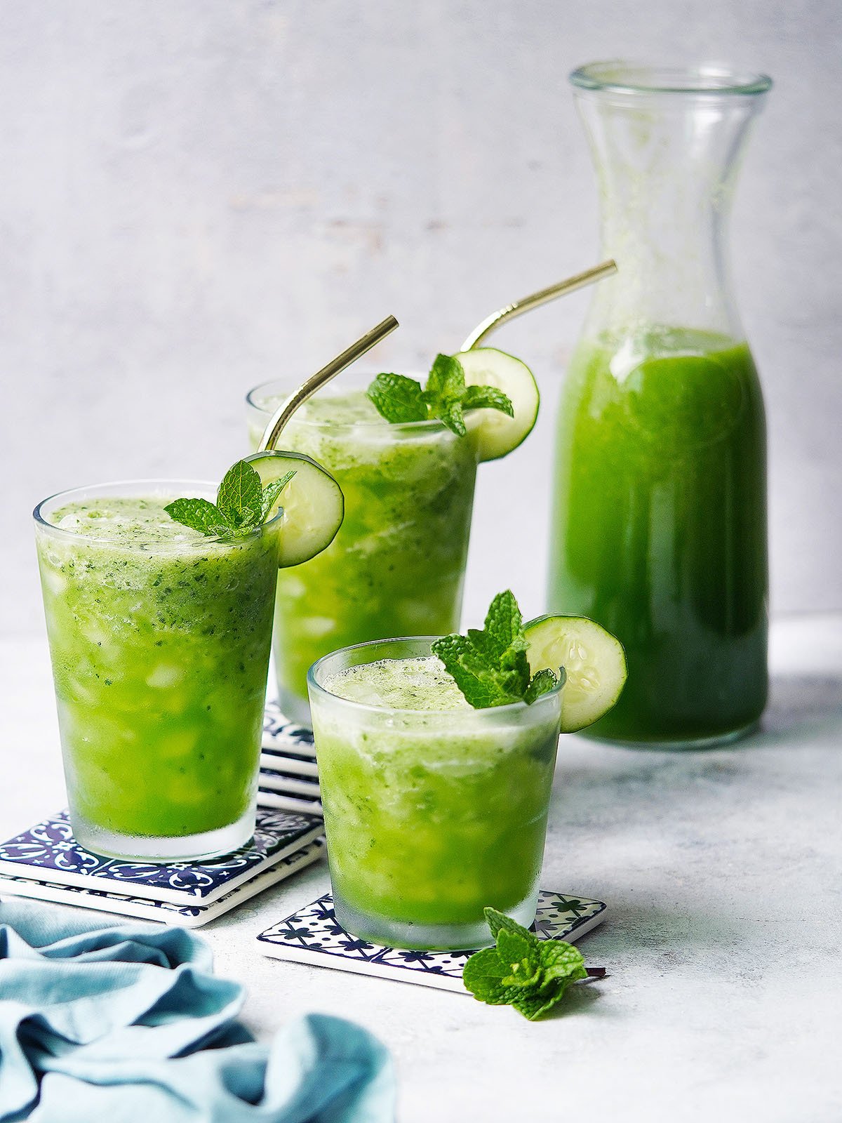 A jar and 3 glasses with Agua De Pepino garnished with a slice of cucumber and mint