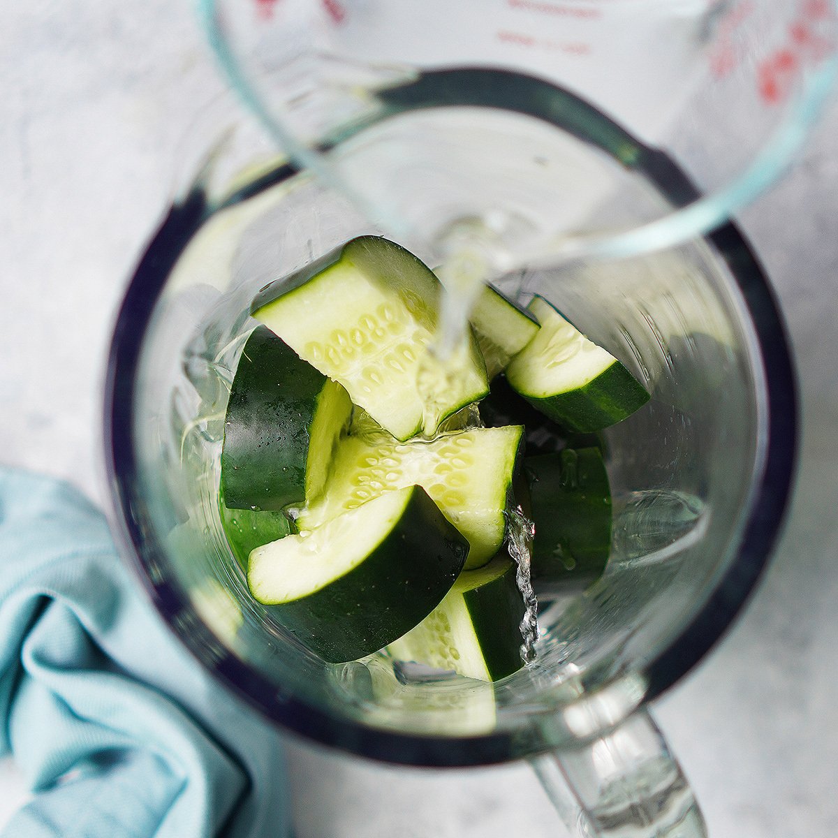 Cucumber slices in a blender glass with water being poured over it.