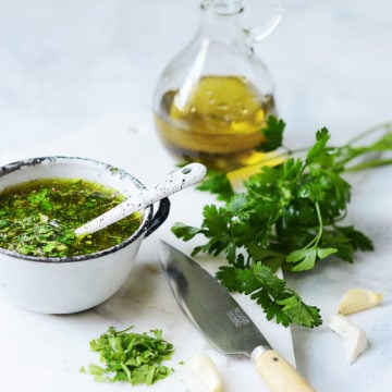 A cutting board with garlic, parsley, a knife and a cup with chimichurri
