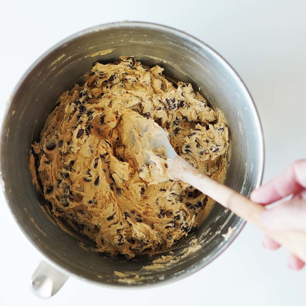A bowl of a mixer with cookie dough and a hand stirring it.
