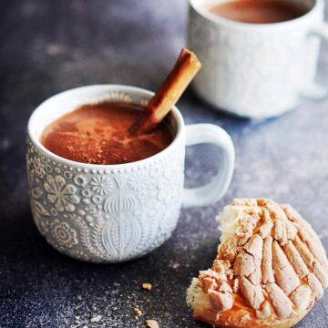 Two blue mugs with hot chocolate garnished with a cinnamon stick and pan dulce on the side.