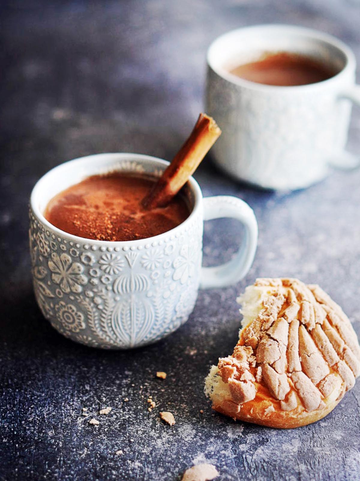 Two blue mugs with hot chocolate garnished with a cinnamon stick and pan dulce on the side. 