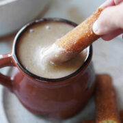 A brown ceramic mug with Atole De Pinole and a hand dipping a churro on the drink.