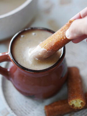 A brown ceramic mug with Atole De Pinole and a hand dipping a churro on the drink.