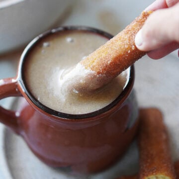 A brown ceramic mug with Atole De Pinole and a hand dipping a churro on the drink.