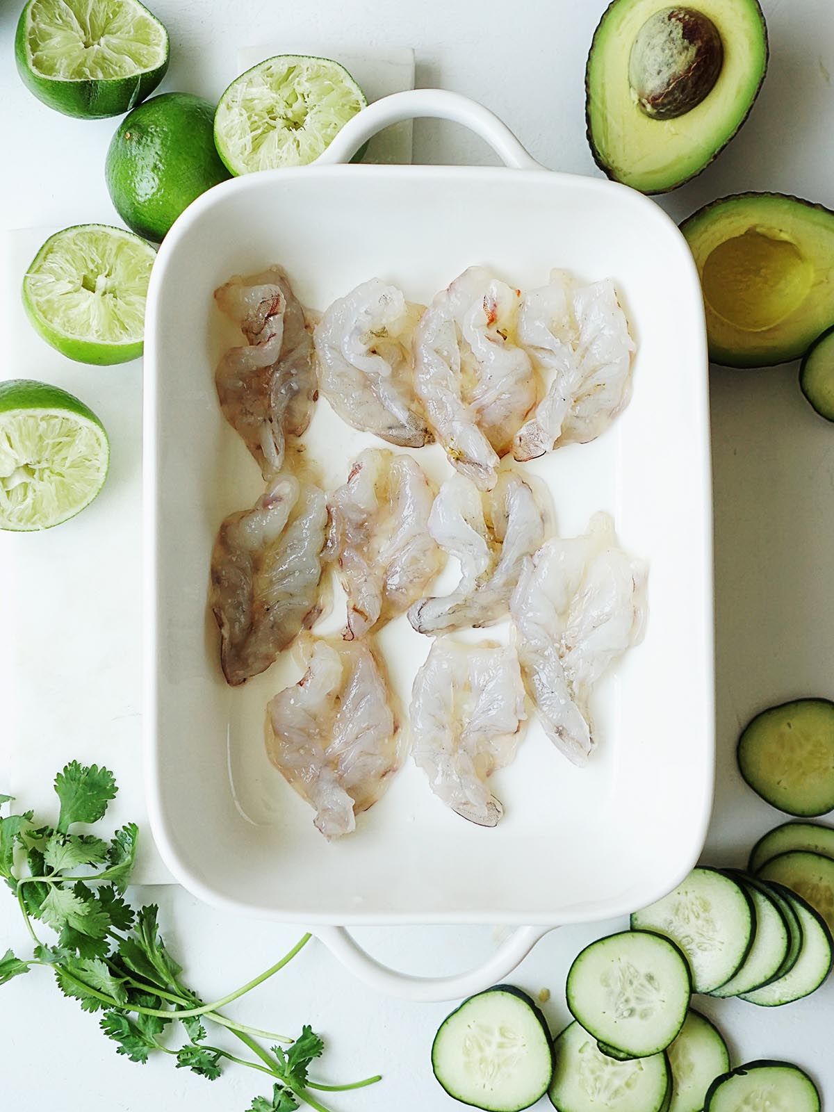 A white dish with raw peeled shrimp and cucumbers on the side as props.