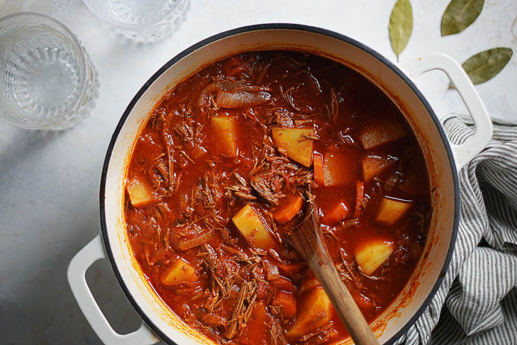 A white pot with shredded beef and potatoes in a red sauce.