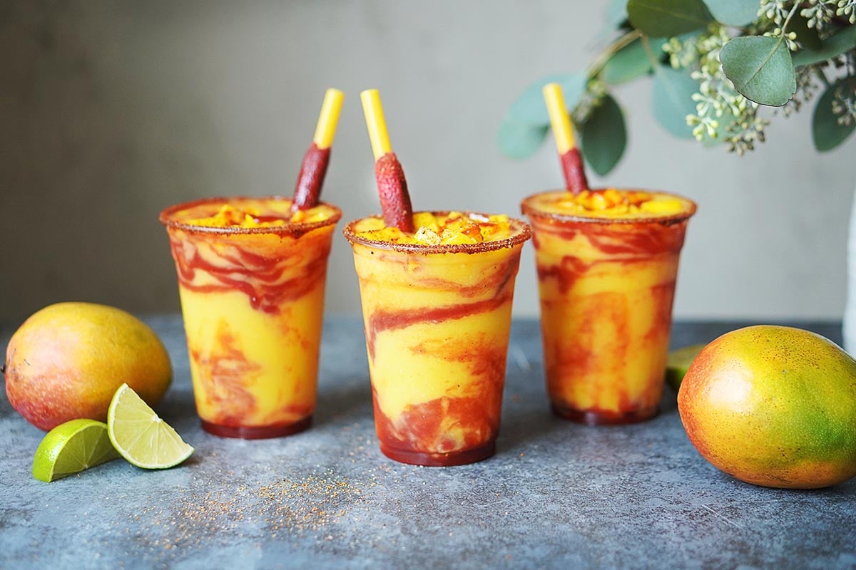 Three plastic cups with mango drink garnished with Mexican candy.