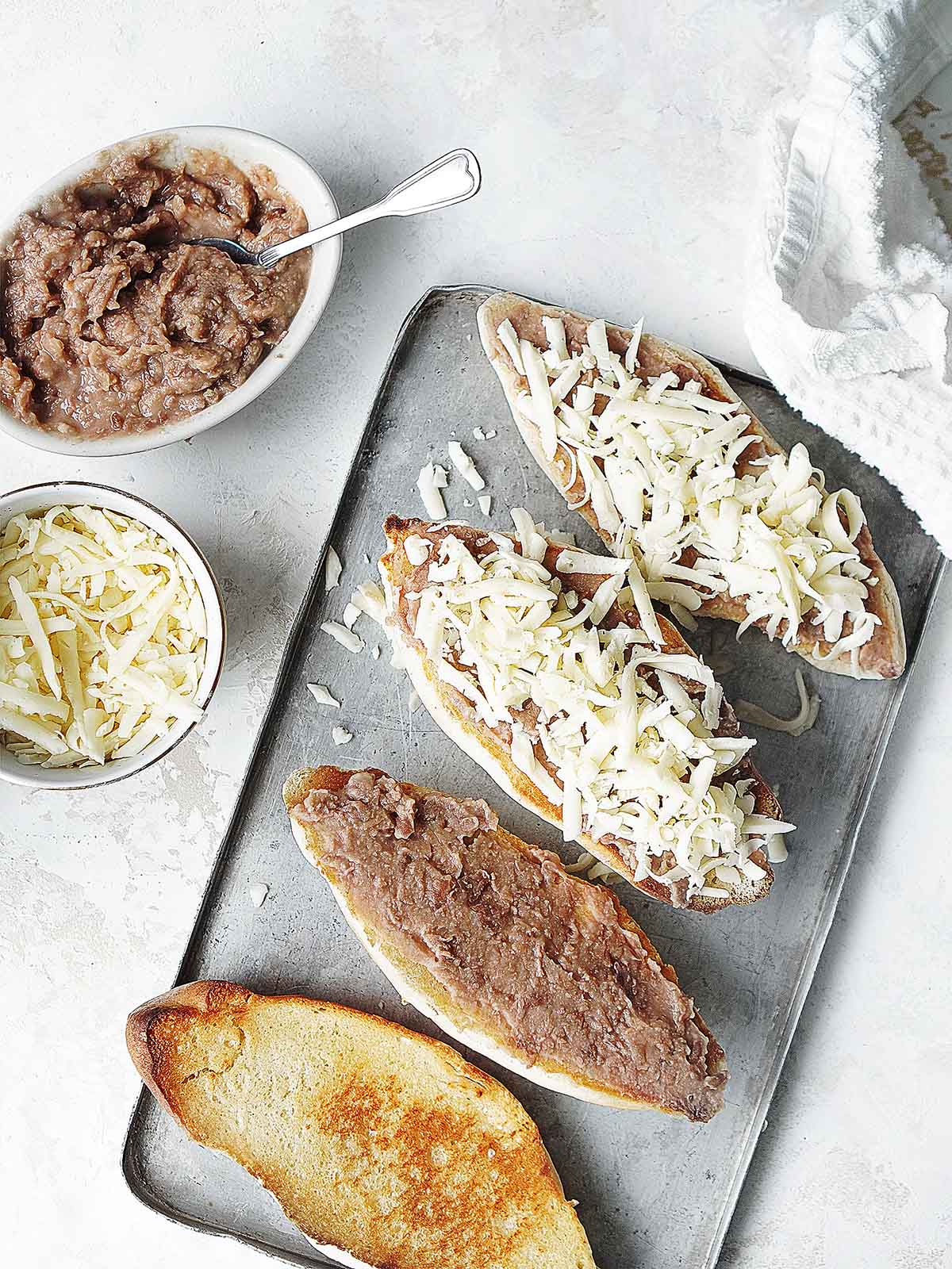 A baking tray with sliced bolillos topped with beans and shredded cheese.