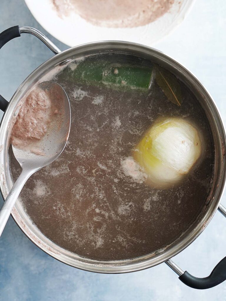 A large pot with beef broth and an onion with a spoon skimming the top of the broth.