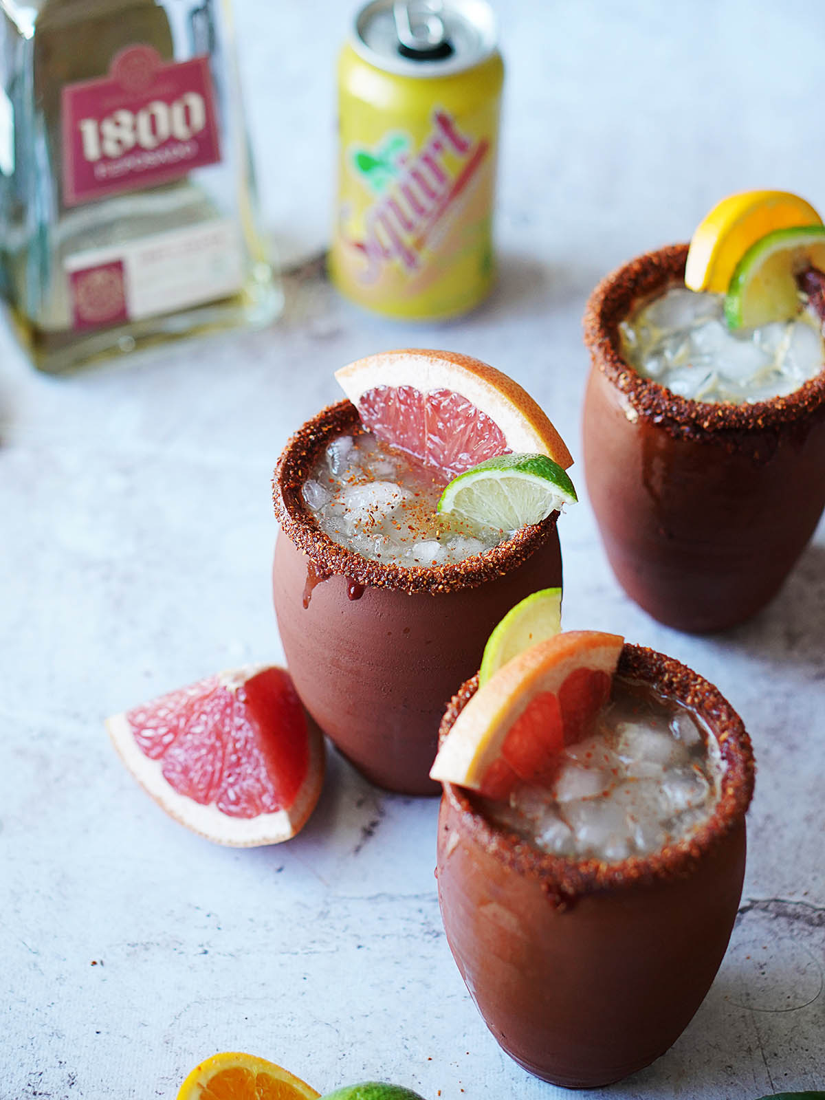 Three cantarito clay cups with filled with a cocktail and topped with a slice of grapefruit and lime with a bottle of tequila in the background.