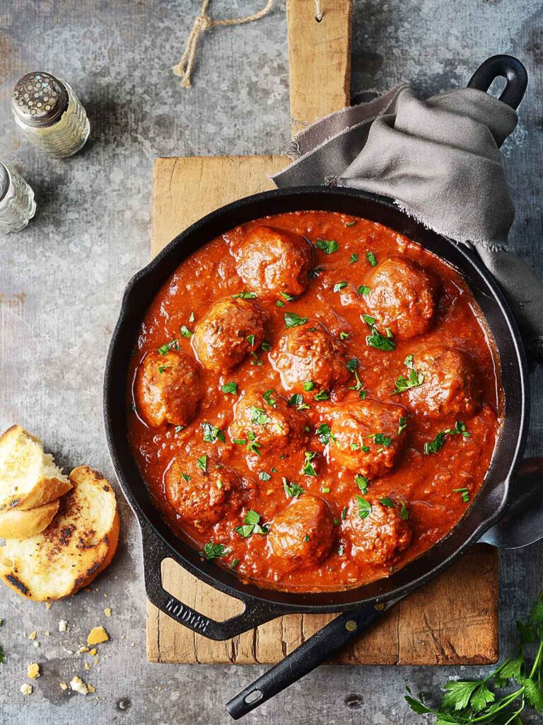Beef meatballs in a black iron skillet with a spoon on the side and some plates.