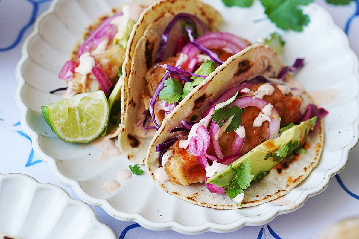 Three Baja fish tacos topped with onions, cabbage and chipotle crema. A slice of lime on the side.