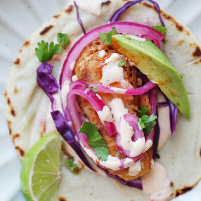 A baja fish taco topped with onions, cabbage and chipotle crema. A slice of lime on the side.