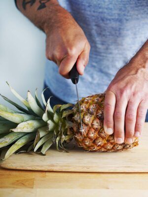 A hand with a knife cutting the top leaves of a pineapple.
