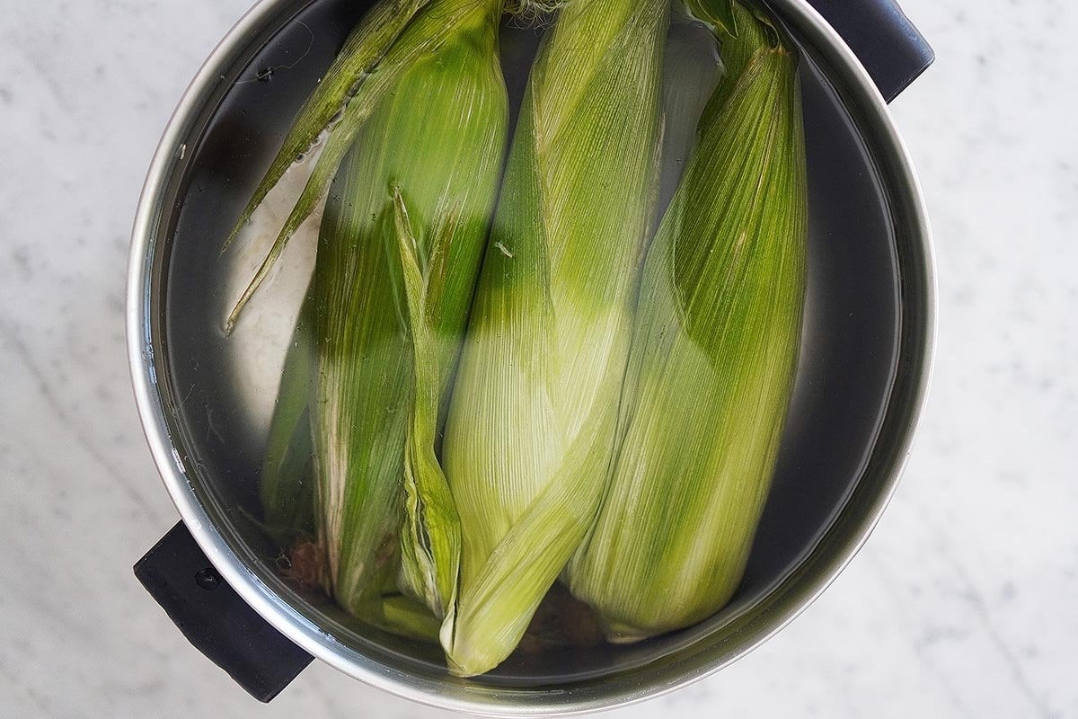 A large pot filled with water and raw corn on the cobs with the husk on. 