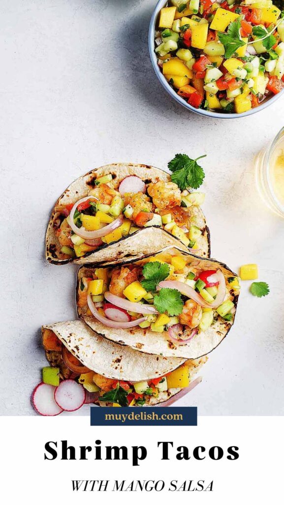 Three shrimp tacos topped with mango salsa on a white board.