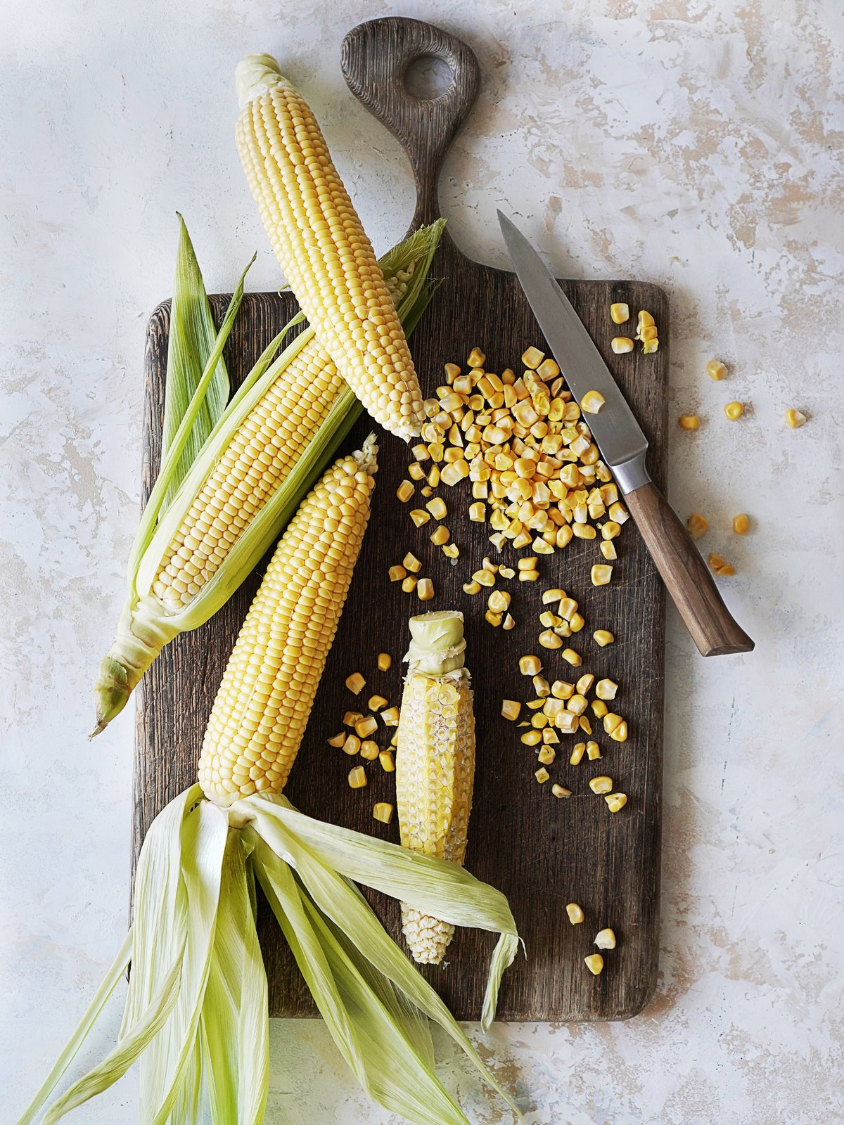 A pretty cutting board with whole corn on the cobs and a knife next to them.