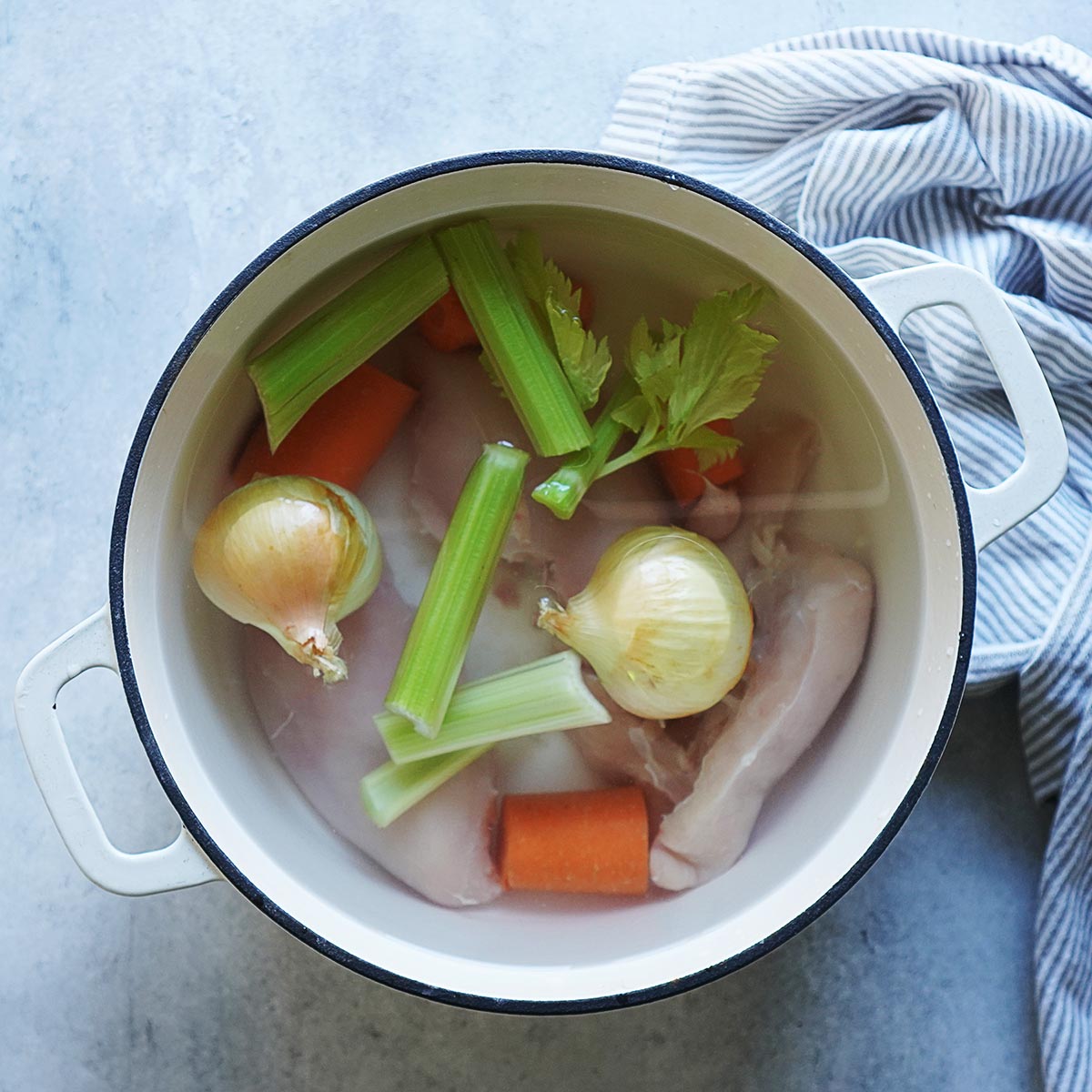 A white stock pot with raw chicken breast, celery, carrots and onions covered in water.