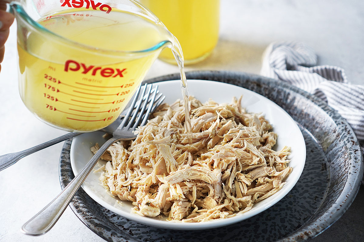 Pouring chicken broth from a measuring cup into shredded chicken on a white bowl.