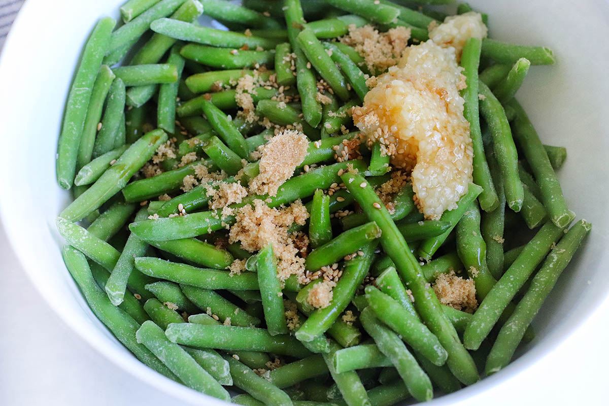 Frozen green beans in a bowl with chopped garlic, brown sugar & sesame seeds.