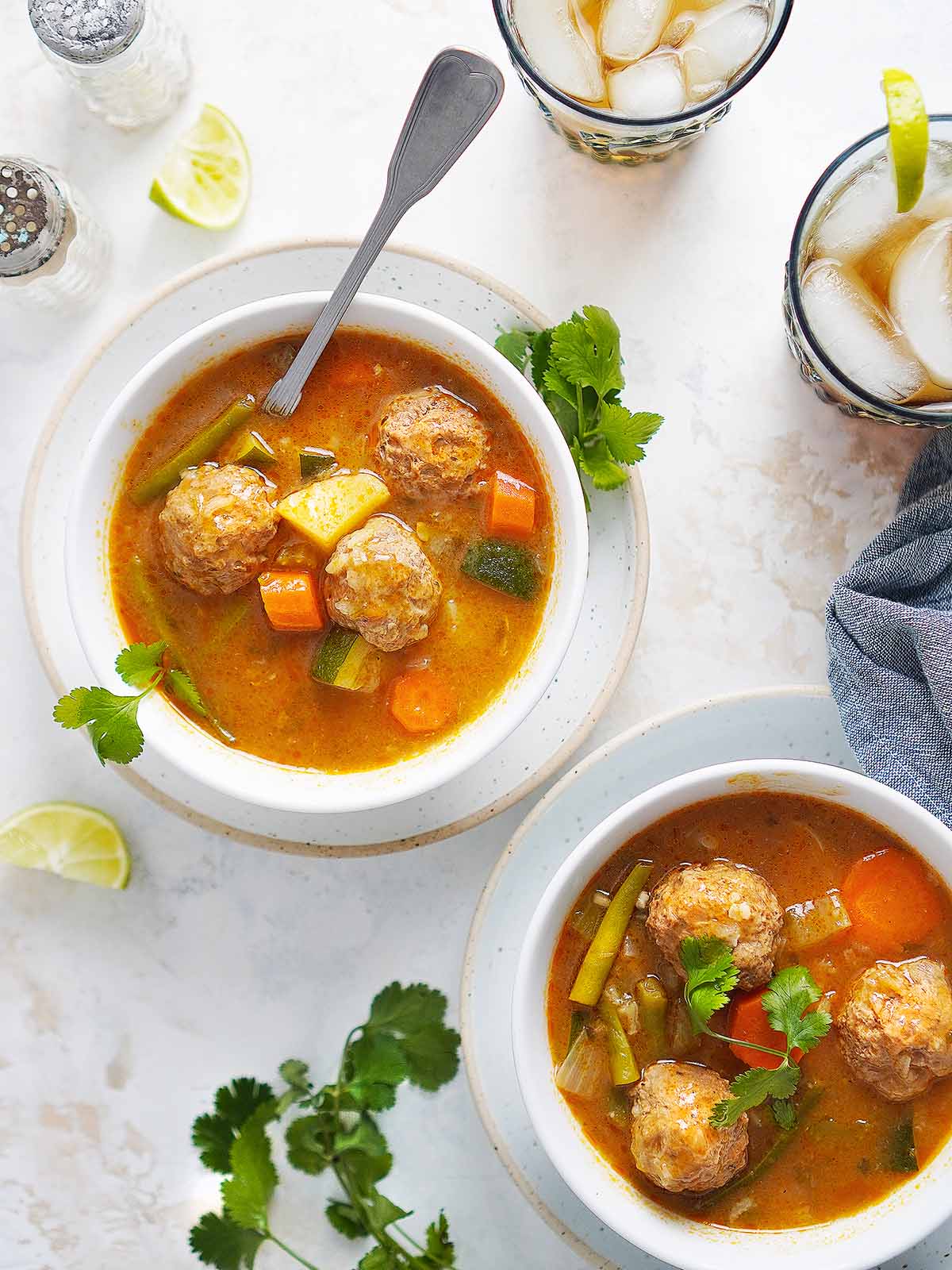Two white bowls with albondigas soup with a spoon and two glasses with drinks on the side.