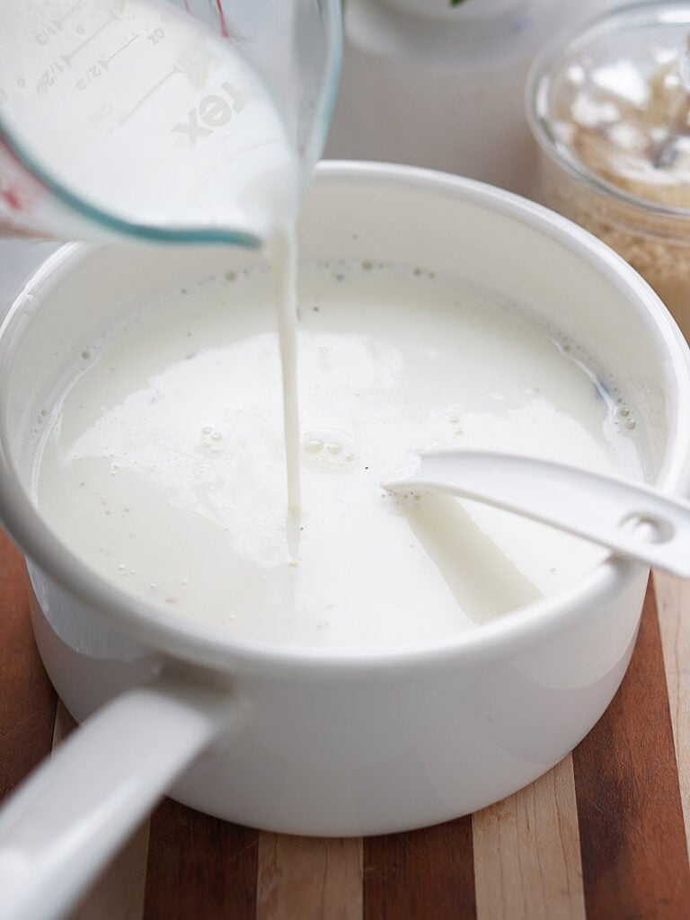 A white saucepan with milk and a measuring cup pouring more milk into it.