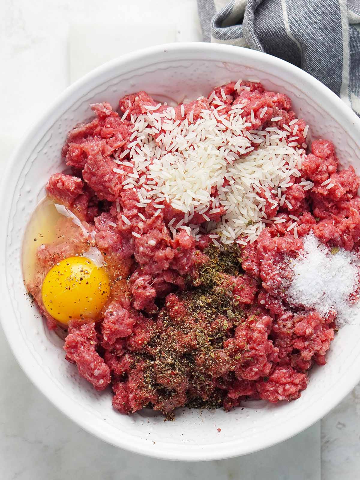 A white bowl with ground beef, oregano, salt, one raw egg and raw rice on top of it.
