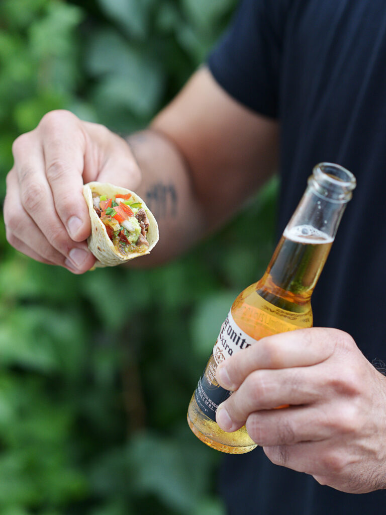 A man holding a taco with one hand and a corona beer with the other hand.