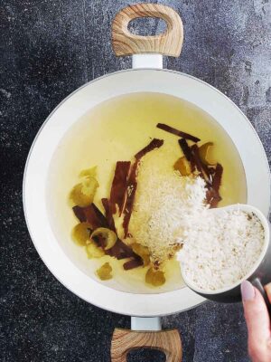 A white saucepan with water, cinnamon sticks and lime peels plus a hand adding rice to it.