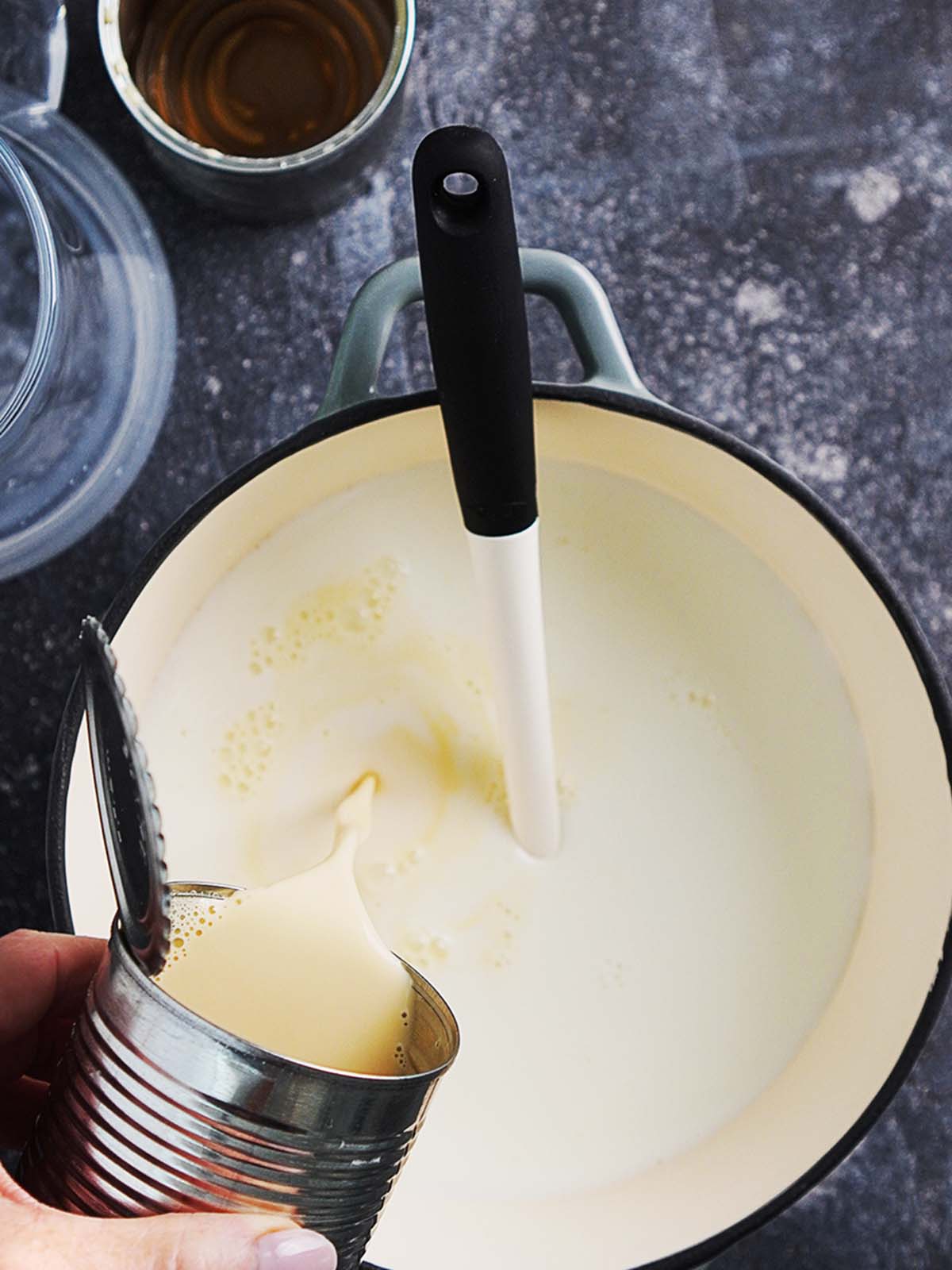 A saucepan with milk and a hand adding condensed milk to it.
