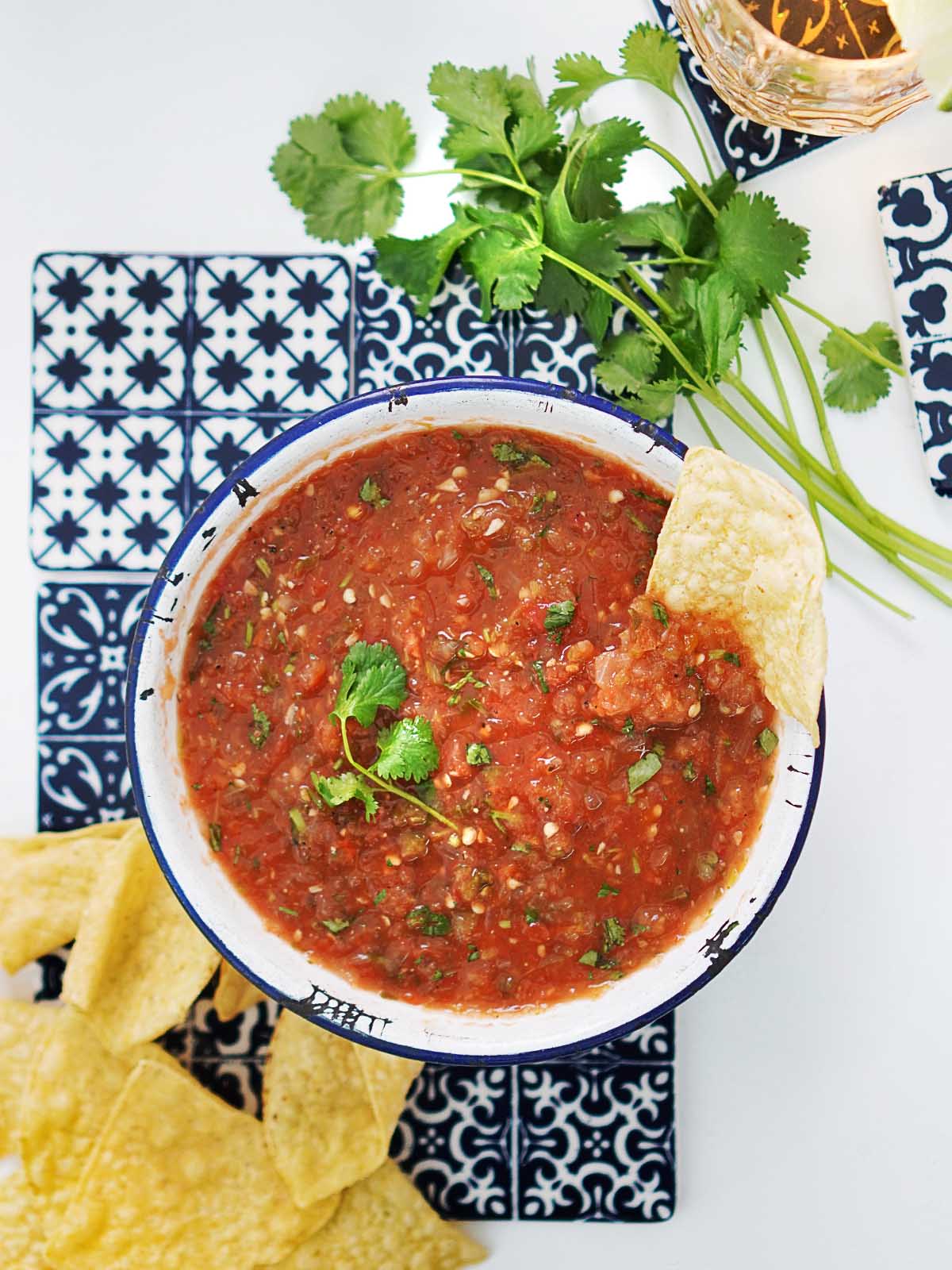 A bowl of red salsa placed on top of blue decorative tiles and chips and cilantro on the side.