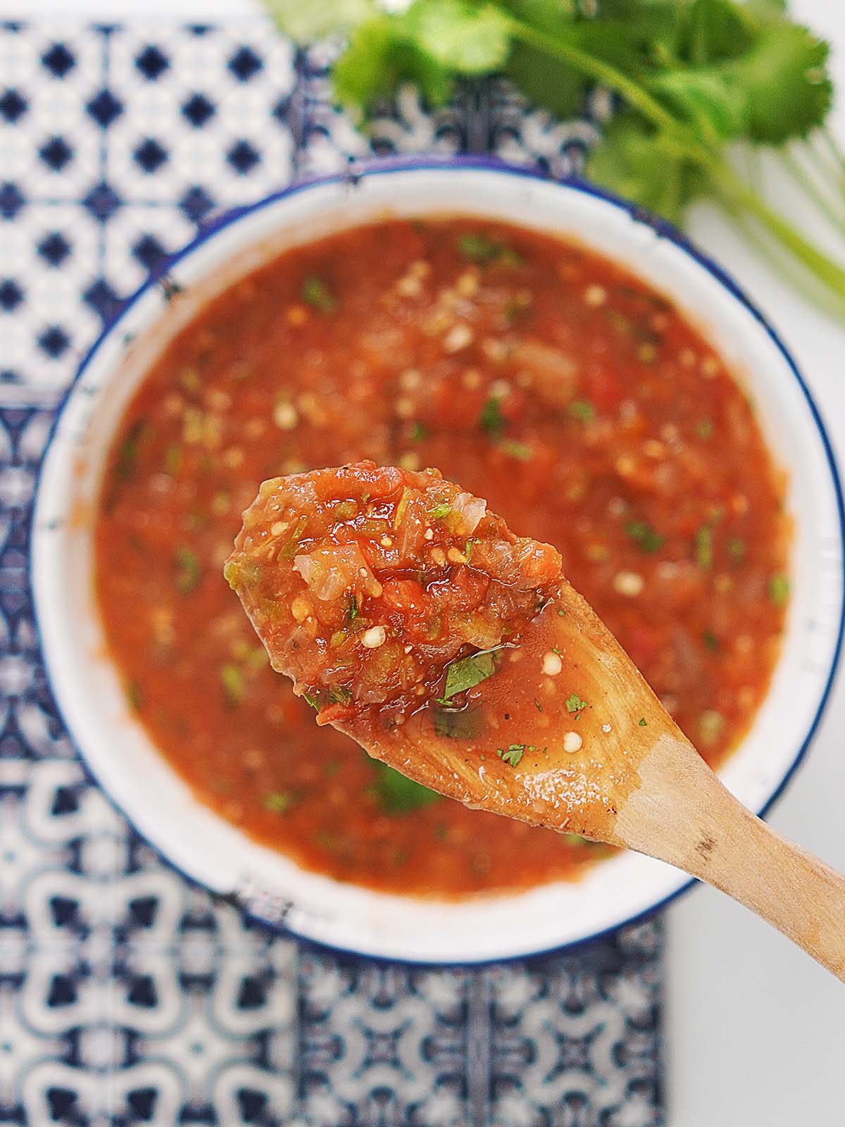 A bowl of red salsa placed on top of blue decorative tiles with a wood spoon. .