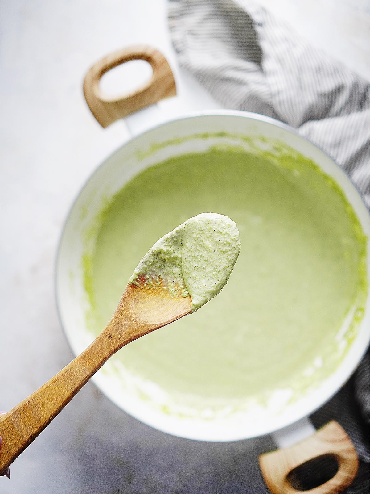 A wooden spoon with a green creamy sauce and a saucepan in the background.