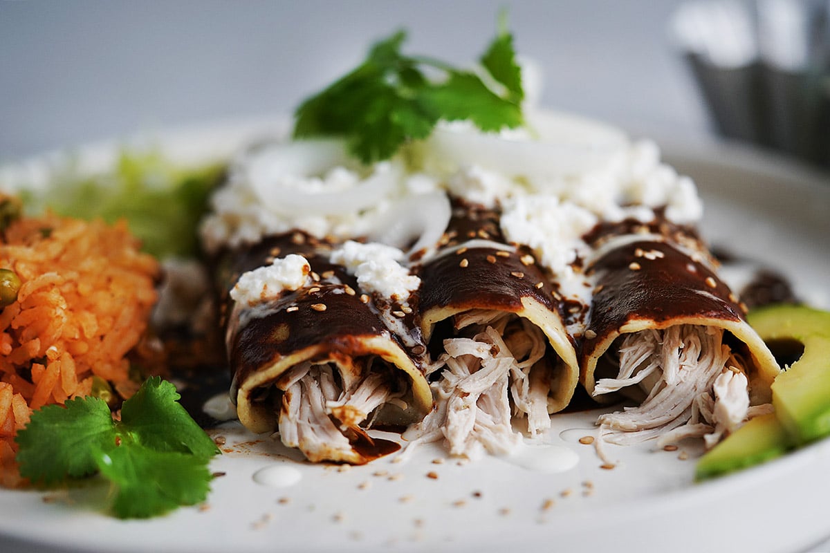 Three mole enchiladas on a white plate topped with sesame seeds, queso fresco and onion slices and Mexican red rice on the side.
