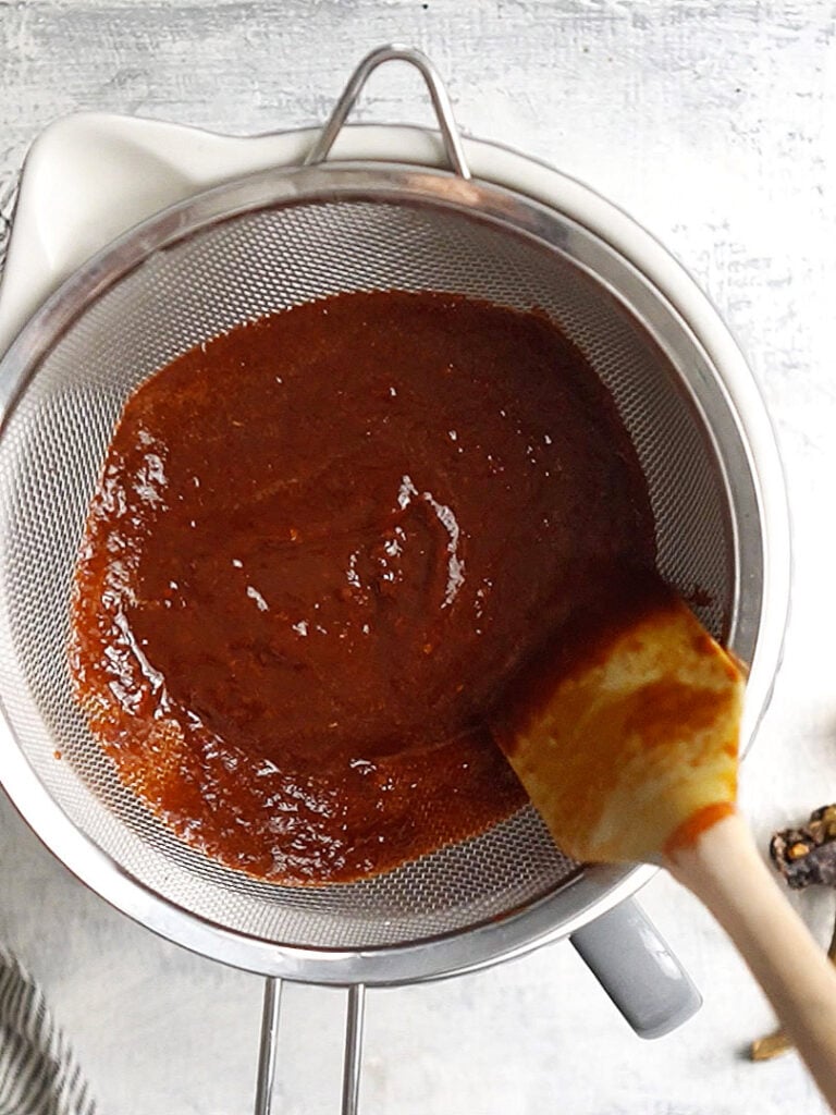 A mesh strainer with red sauce and a spatula pushing it thru.