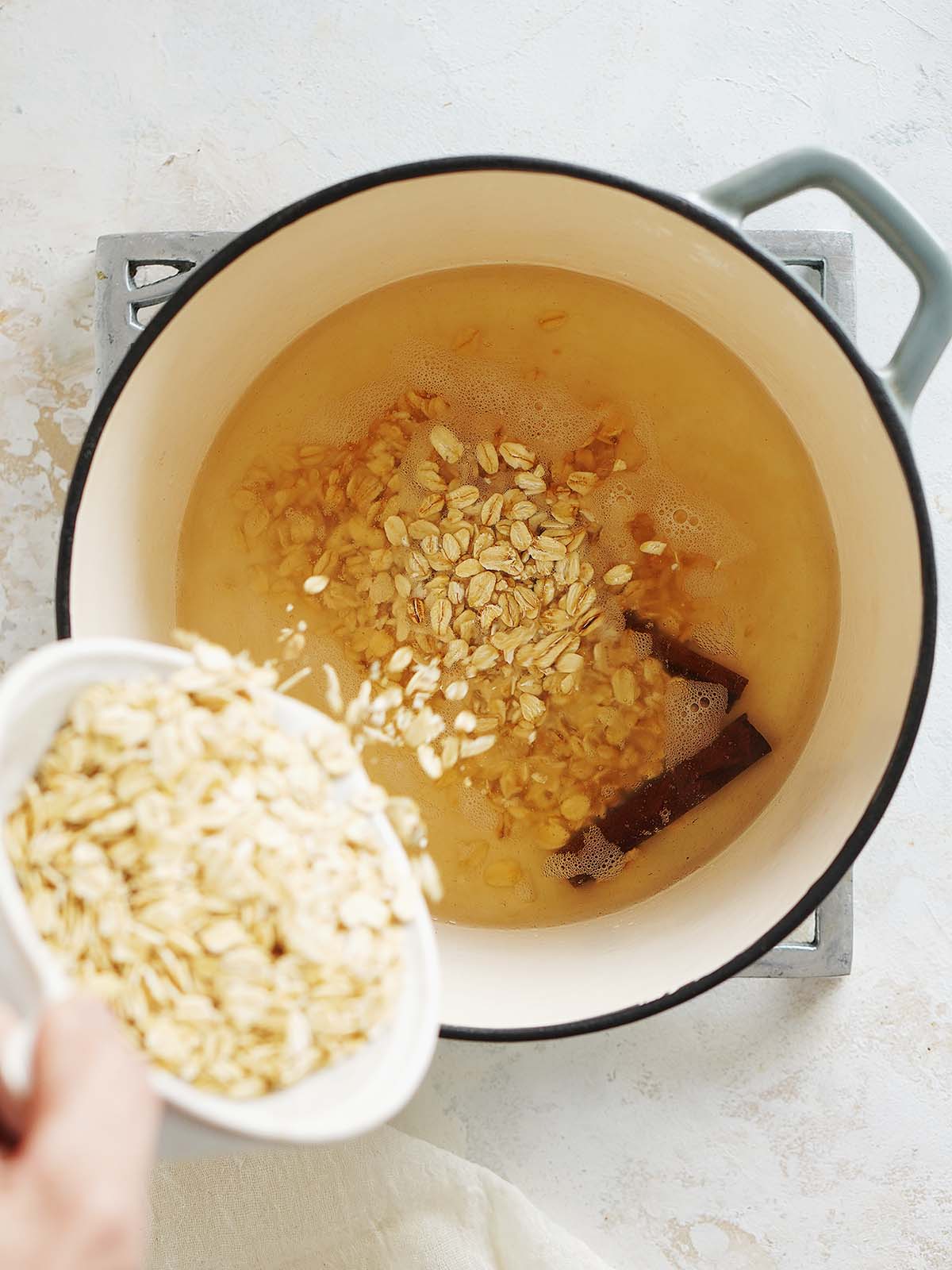 A medium pot with water, cinnamon sticks and pouring oatmeal flakes in it.