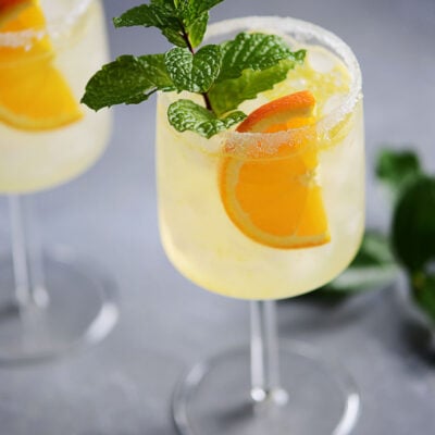 Two cocktail glasses with a clear drink garnished with an orange slice and a mint sprig.