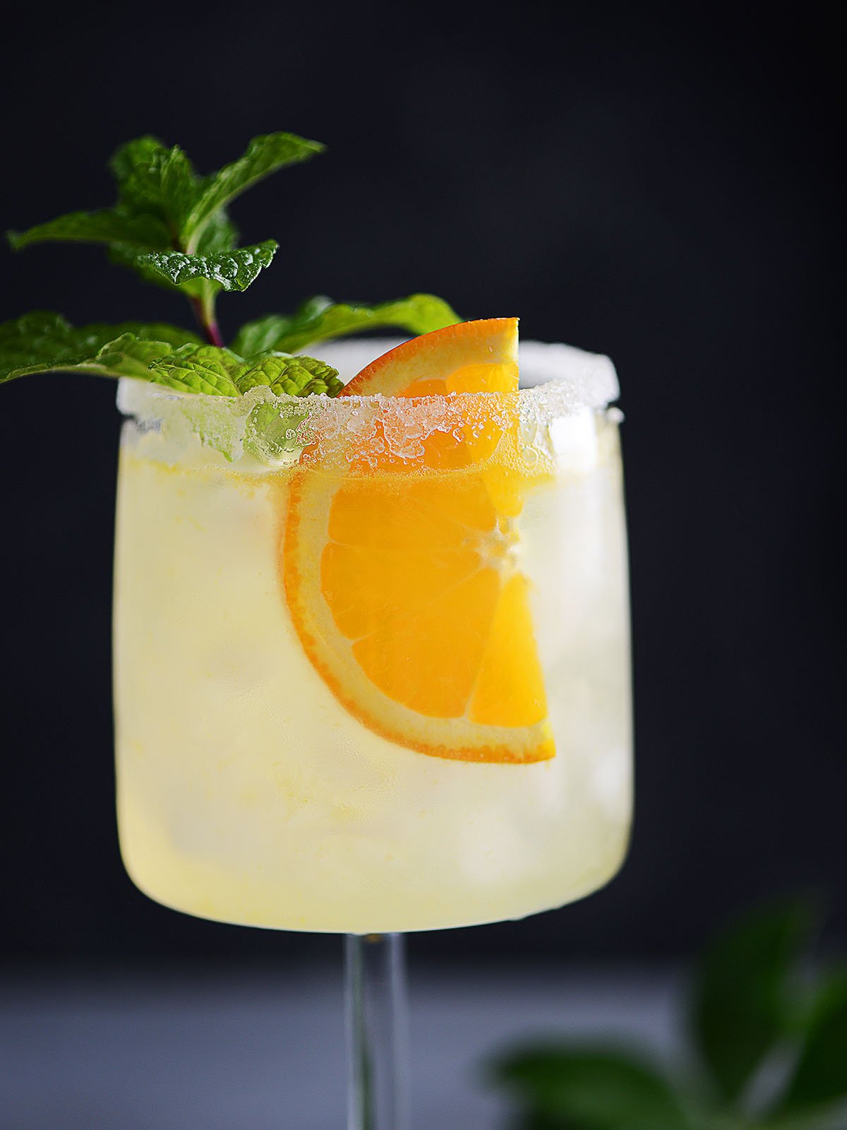 A cocktail glass with a clear drink garnished with an orange slice and a mint sprig.